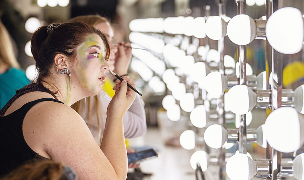 Actress adjusting her makeup in front of a long mirror with bright lights