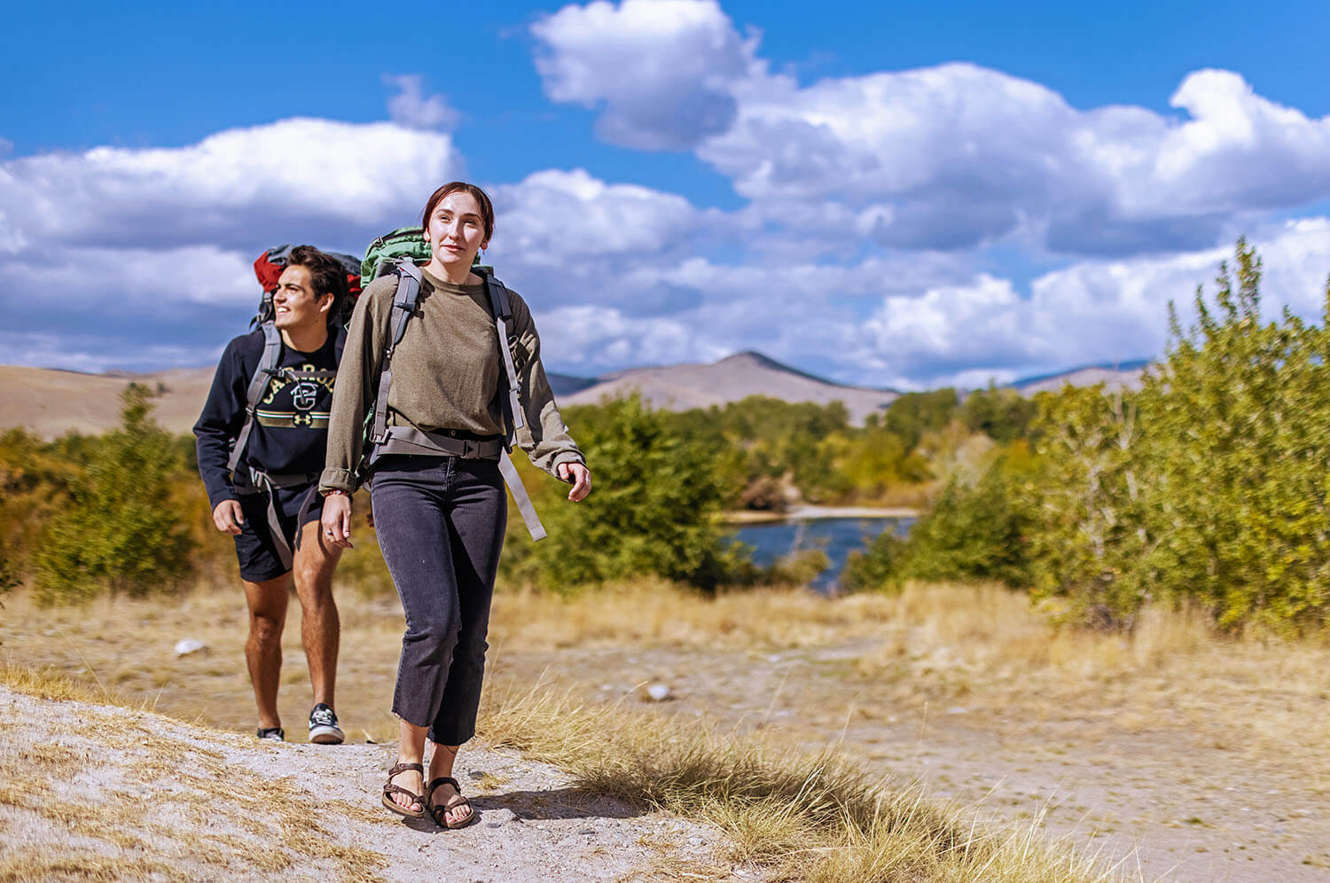 Two students on a dirt trail while wearing hiking backpacks and large mountains in the distance