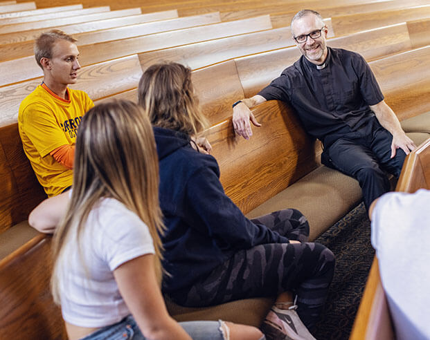 Minister sitting in a pew chatting with a group of students
