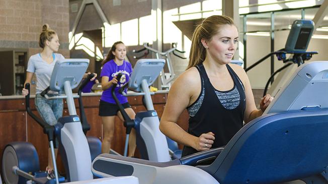 Student starting their workout on a treadmill, two other students in the background on elipticals